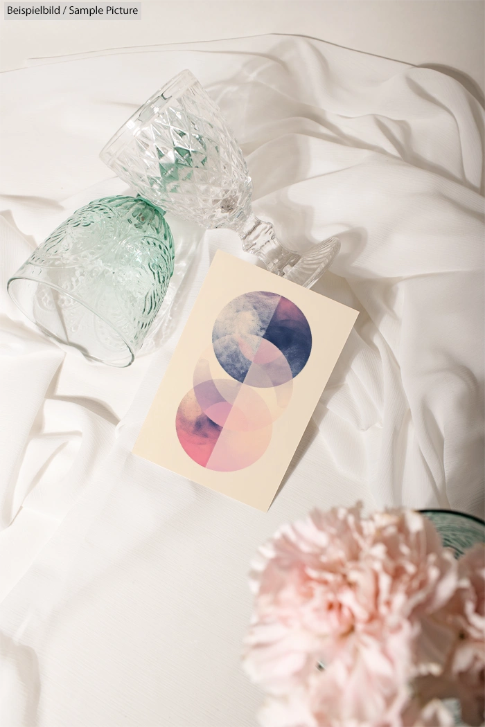 Abstract geometric card with pink and blue circles on fabric, surrounded by upside-down glassware and flowers.