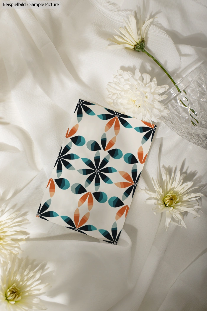 Geometric patterned notebook on white fabric surrounded by white flowers and crystal vase.