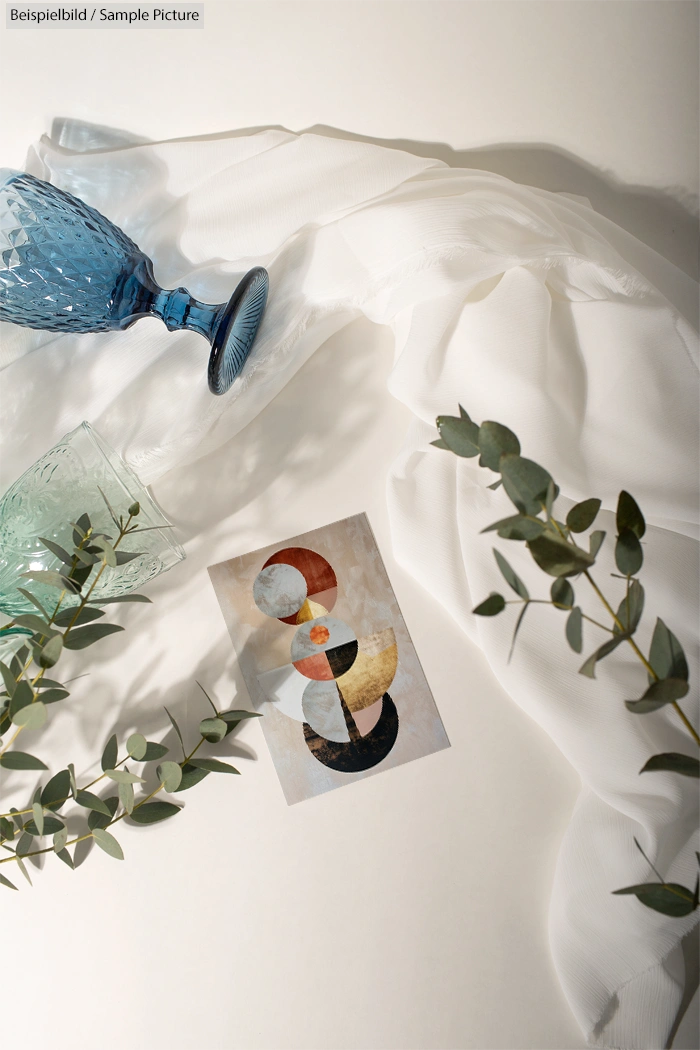 Abstract painting with geometric shapes on paper, surrounded by draped fabric, glass cup, and eucalyptus leaves.