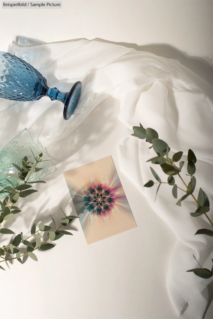 Artistic flat lay with glassware, white cloth, eucalyptus sprigs, and abstract art on a light background.