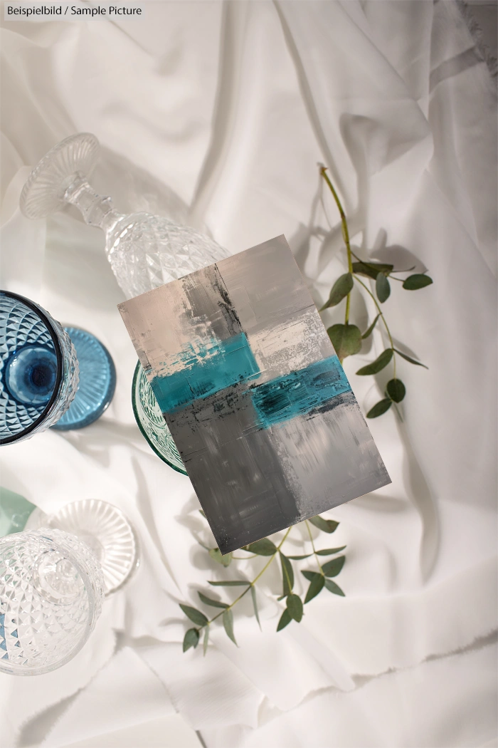 Abstract painting with blue, teal, and gray blocks on draped white fabric, surrounded by glassware and green leaves.