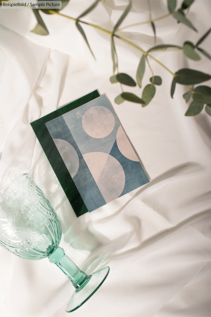 Decorative green goblet and geometric notebook on white cloth with eucalyptus leaves.