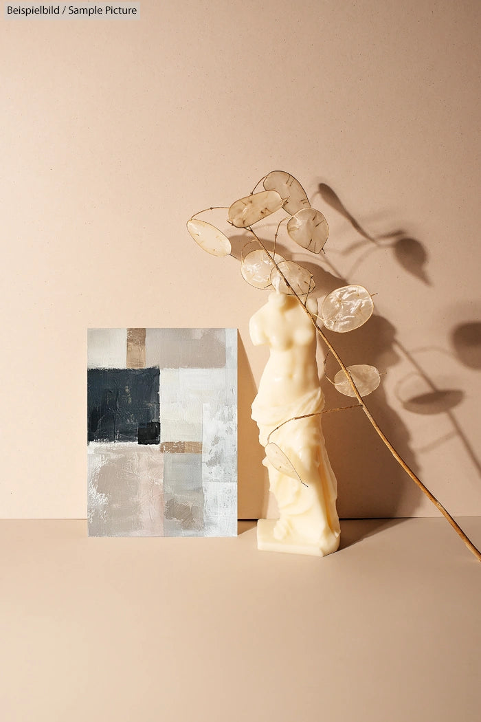 Abstract beige and black art piece beside an ivory statuette and a dried branch casting shadows on a neutral background.