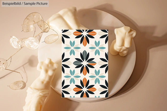 Decorative card with geometric floral pattern in black, blue, and orange, placed on a plate with sculptural candles.