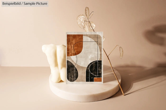 Abstract art print with curved shapes in earth tones, set on a white stand with dried plants against a beige background.