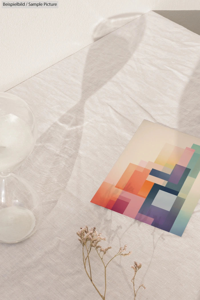 Abstract geometric art print on textured table with dried flowers, alongside a translucent hourglass casting a shadow.