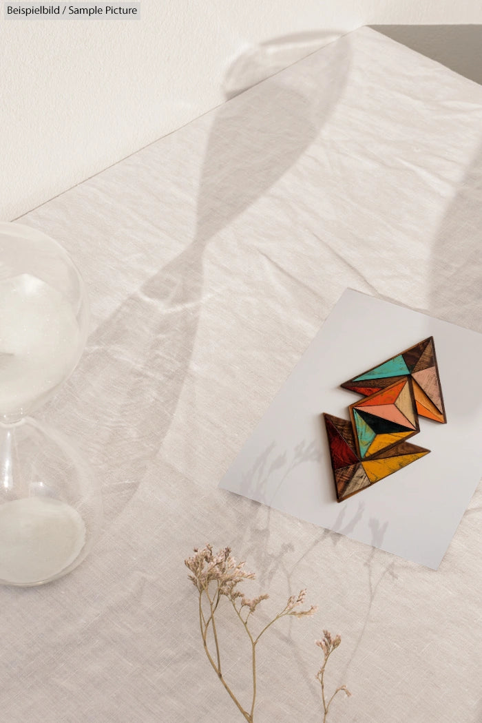 A minimalist scene with a geometric artwork, dried flowers, and a shadowed hourglass on a textured surface.