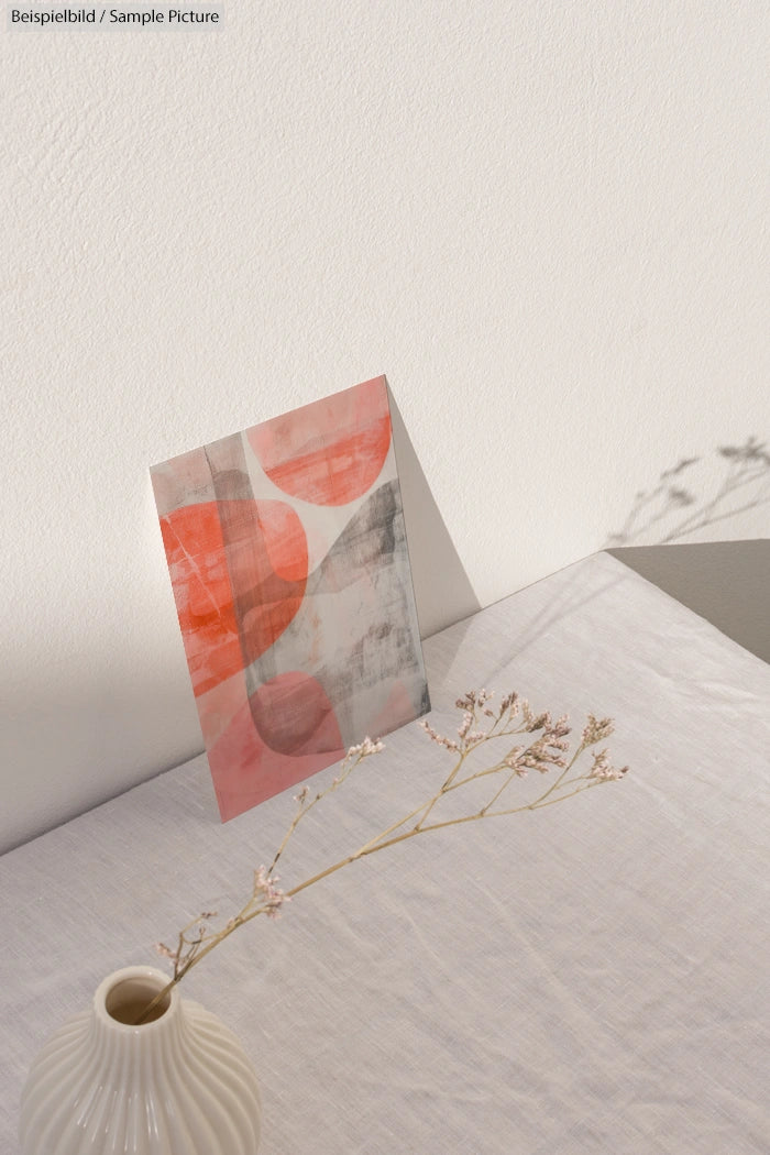 Abstract art print in red and gray tones leaning against a wall, on a table with a vase holding delicate dried flowers.