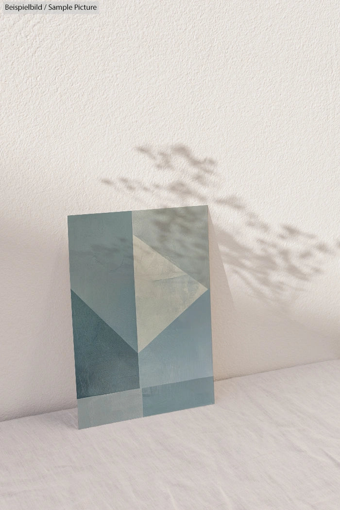 Abstract geometric art piece in shades of blue and green against a textured white wall with shadow patterns.