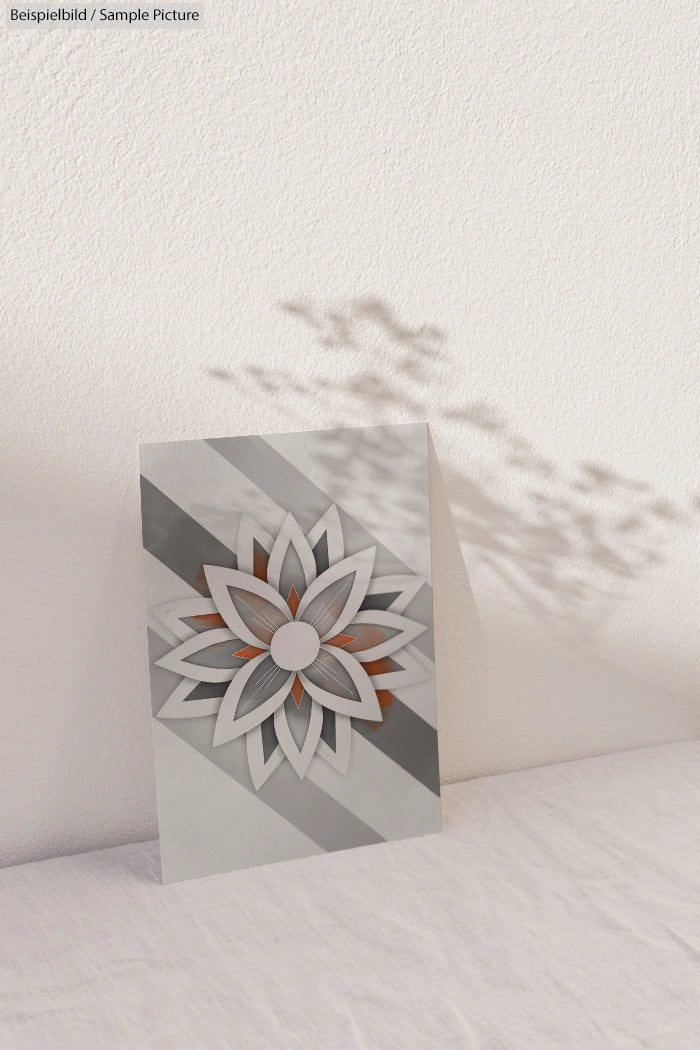 Geometric floral card with gray and orange tones on a white surface, casting artistic shadows on a textured wall.