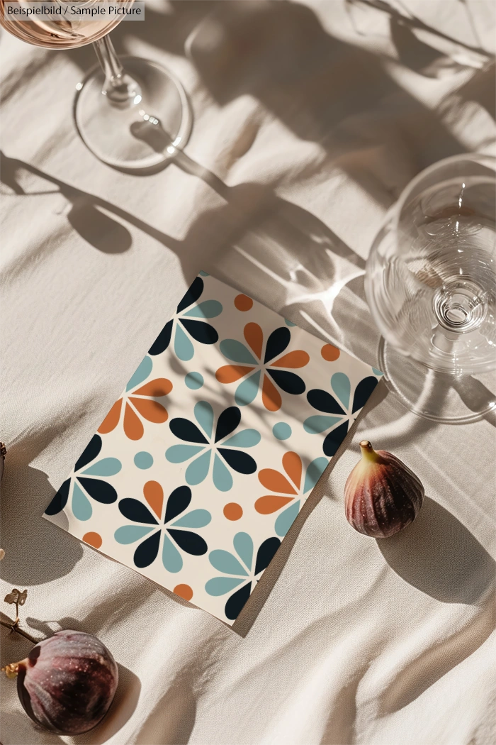 Patterned paper with retro floral design on white fabric, surrounded by figs, two wine glasses, and diagonal shadows.