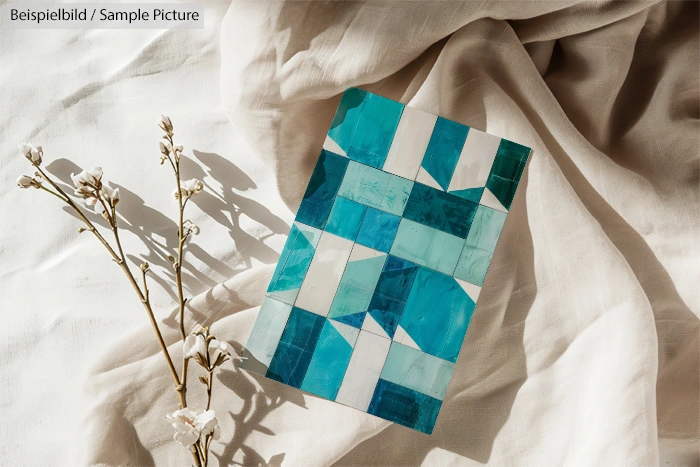 Geometric artwork with shades of blue and white on a beige fabric, next to a sprig of dried white flowers.