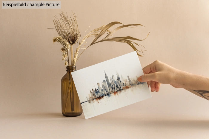 Hand holding cityscape art print next to vase with dried grasses.