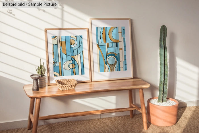 Modern abstract art prints in frames on a wooden bench with a cactus and small plants, sunlight streaming through blinds.