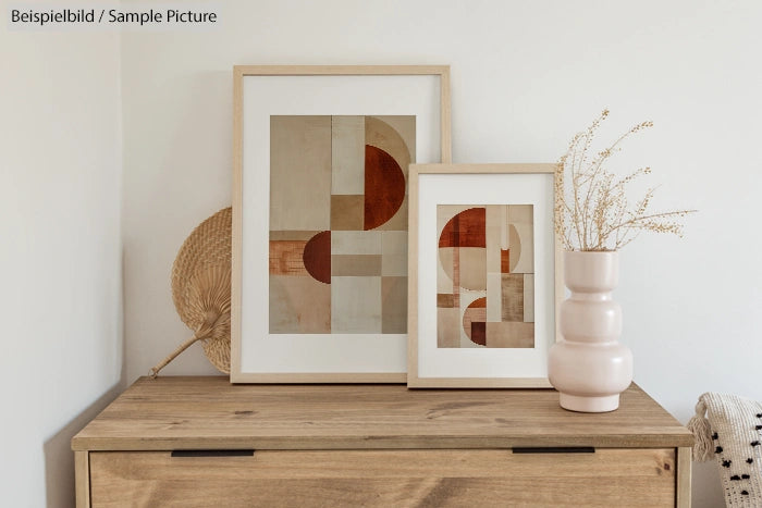 Minimalist decor: two abstract geometric prints in frames on a wooden cabinet, alongside a woven fan and ceramic vase.