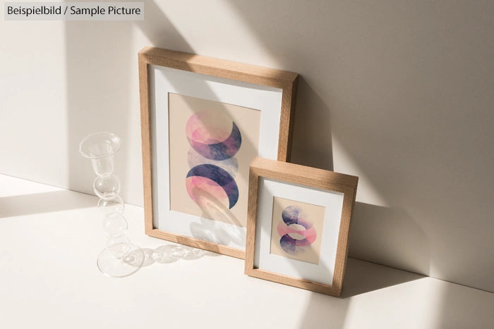 Two framed abstract prints with geometric designs in warm light, beside a sculptural glass object.