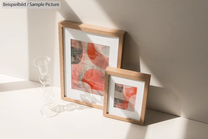 Two abstract paintings with red and gray swirls in wooden frames, placed against a beige wall in sunlight.