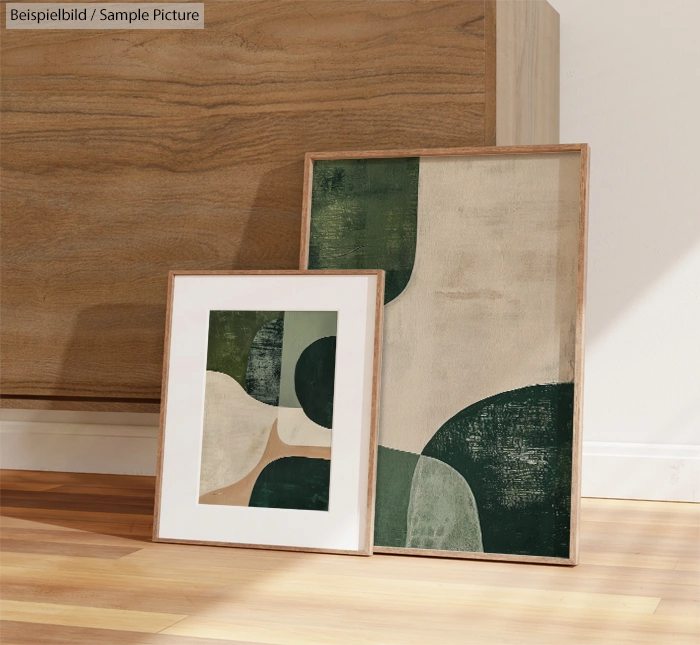 Two framed abstract art prints with green and beige geometric shapes, leaning against a wooden cabinet on a floor.