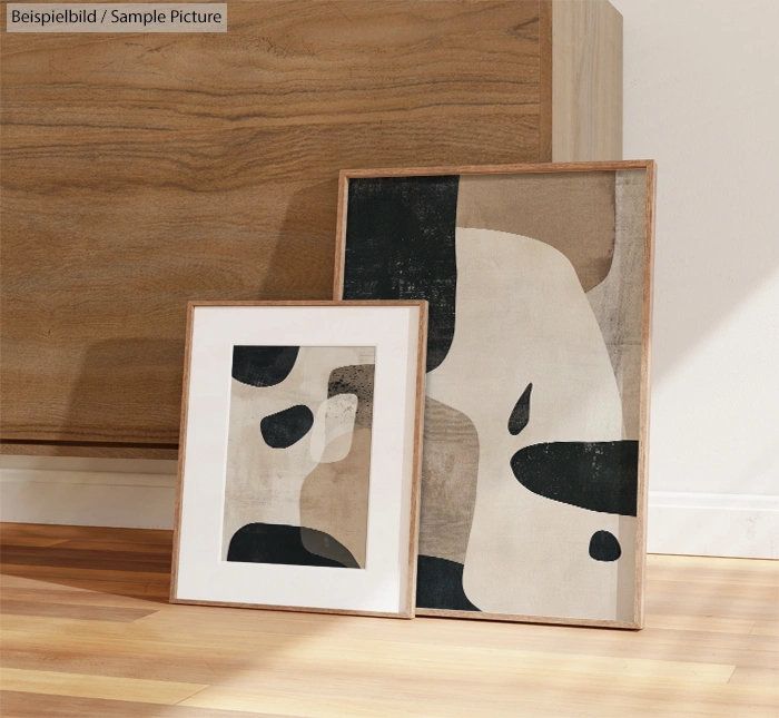 Two abstract paintings with black, beige, and white organic shapes, in wood frames, leaning against a wooden cabinet.