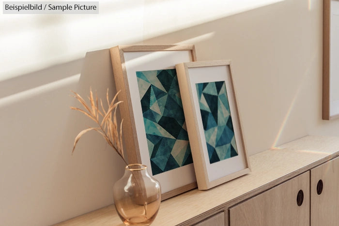 Two abstract geometric art pieces in wooden frames on a shelf, with a dried plant in a clear vase.