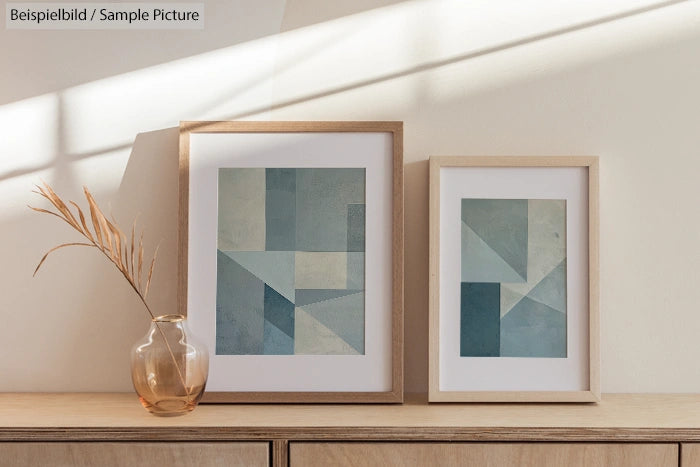 Two framed geometric art prints in pastel colors on a wooden shelf with a vase and grass, sunlit background.