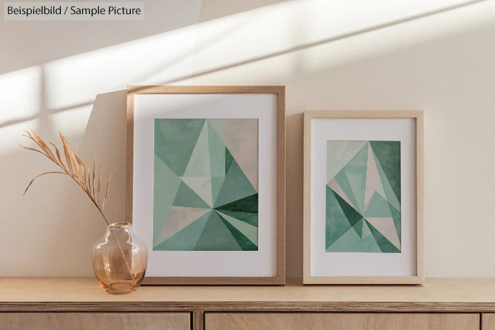 Two framed geometric art prints with green hues on a wooden console next to a glass vase with dried grass.