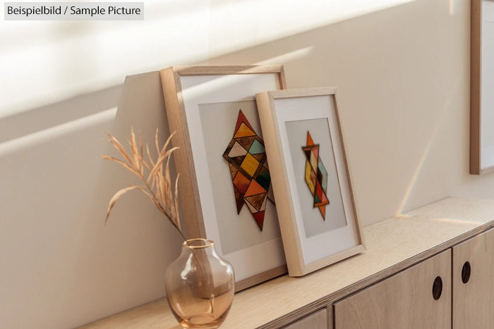 Framed geometric artworks on wooden cabinet with dried plant in glass vase.