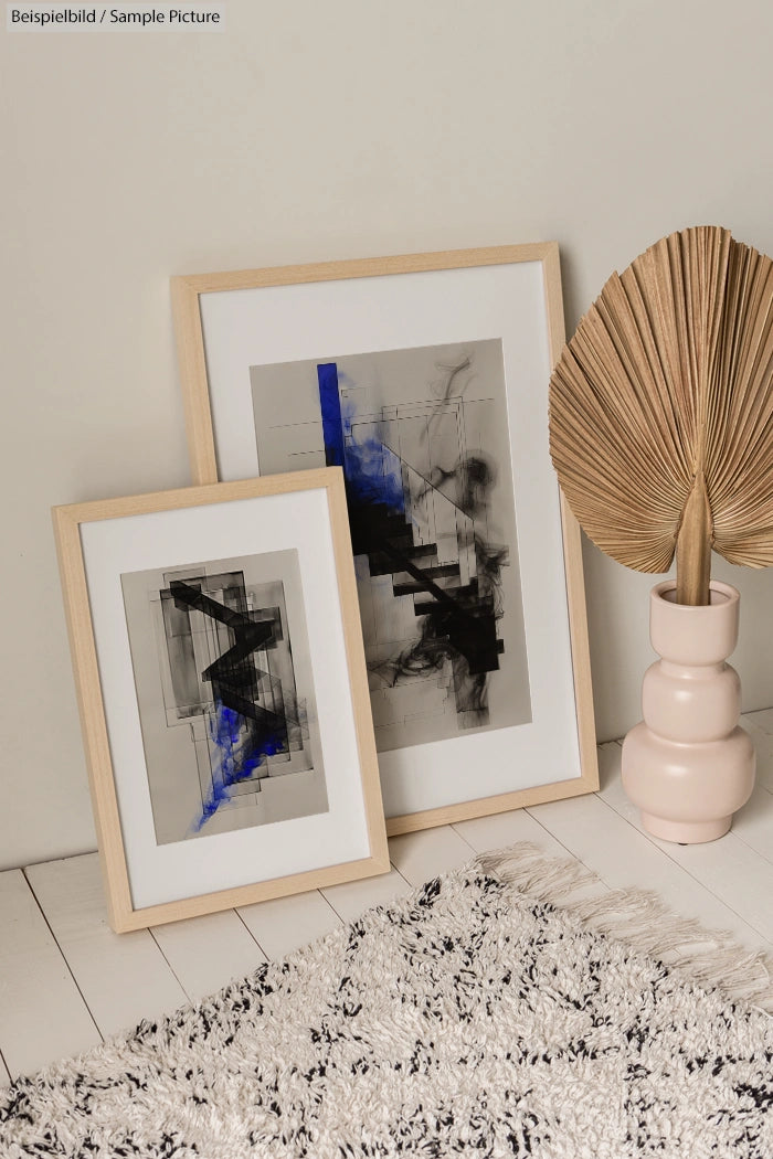 Two framed abstract artworks featuring black and blue geometric patterns, placed on a light floor next to a pink vase with a dried plant.