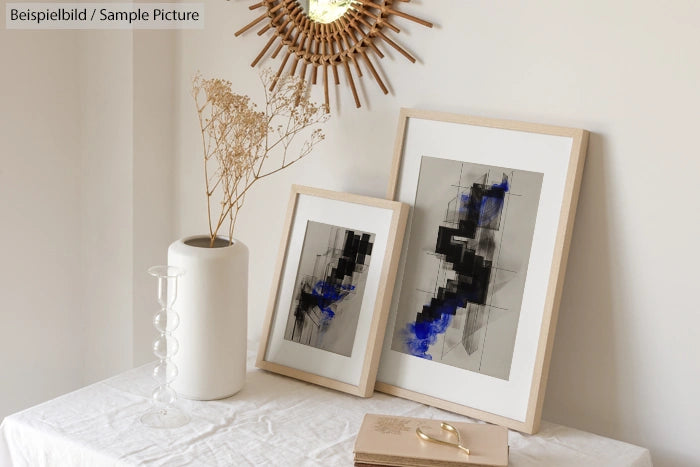 Framed abstract art with black and blue details on a table with a white vase and dried flowers, beneath a sunburst mirror.