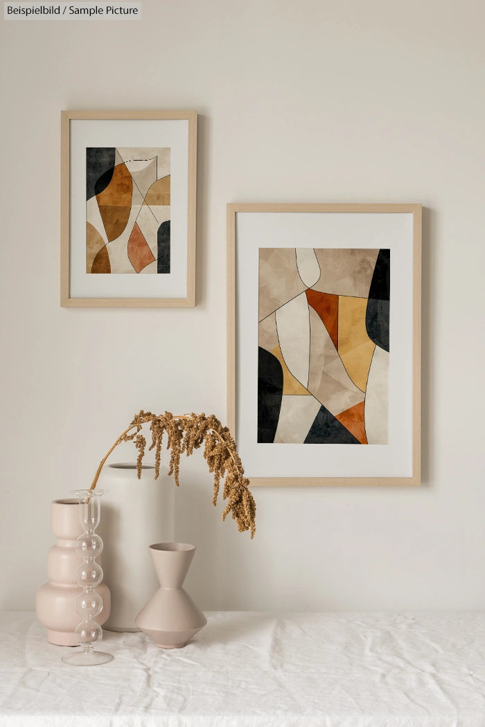 Framed abstract paintings on a beige wall above a table with a decorative vase and dried plant.