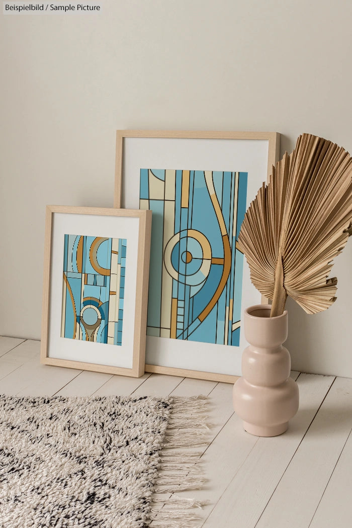 Two framed geometric art prints with blue and beige tones, next to a vase with a dried palm leaf, on a wooden floor.