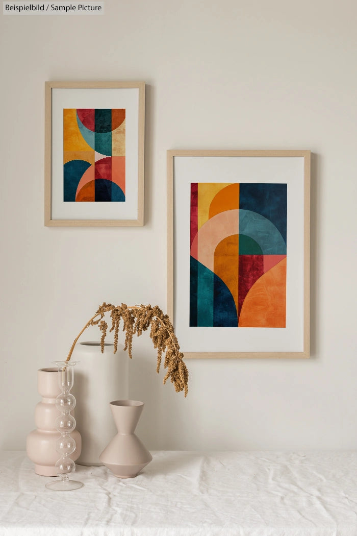 Two framed abstract paintings with colorful geometric shapes hanging on a neutral wall above vases with dried plants.