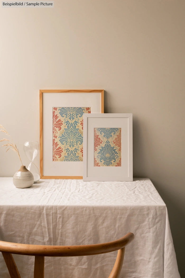 Two framed patterned artworks on a linen-covered table with a wooden chair, hourglass, and dried plant in a vase.