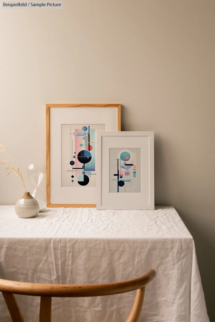 Minimalist room with framed geometric art prints on a table covered with a white cloth, wooden chair in foreground.