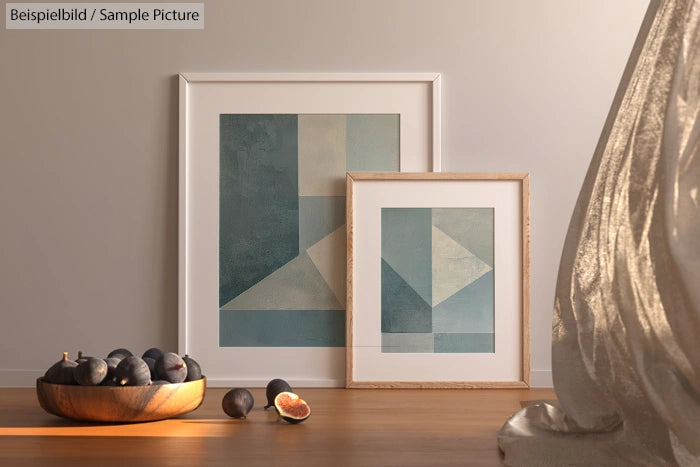 Two framed abstract geometric artworks on floor near bowl of figs and draped fabric. Soft lighting accentuates decor.