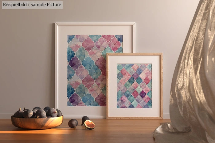 Two framed abstract art pieces with geometric patterns in pink and teal, next to a bowl of figs, on a wooden surface.