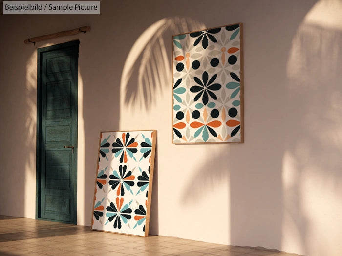 Two geometric pattern artworks leaning against a beige wall with sunlight and shadows, near a closed teal door.