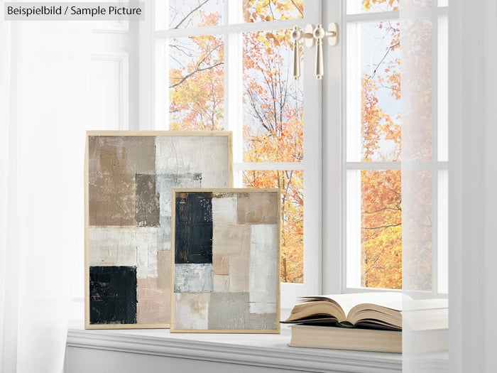 Abstract paintings with neutral tones by a window with autumn leaves outside; open book on sill.