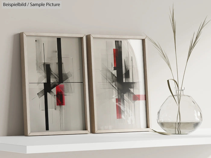 Two abstract art pieces with geometric black and red shapes framed on a shelf beside a vase with dried grass.