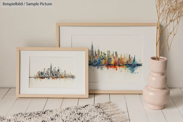 Two framed watercolor cityscape paintings on a white floor next to a beige vase with dried branches.