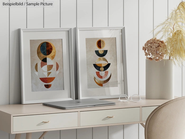 Minimalist desk with laptop, framed geometric art, and dried flowers on a pale wooden surface.