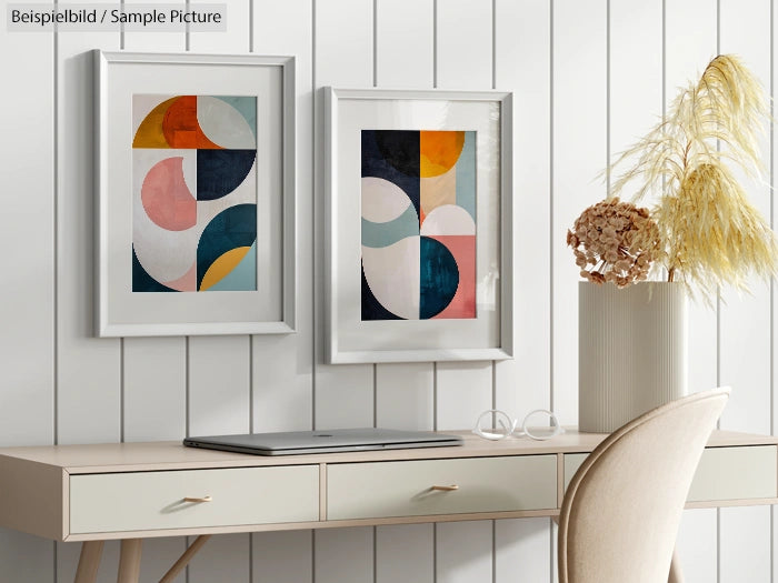 Minimalist workspace with two framed abstract paintings, laptop on desk, pampas grass in vase, and modern chair.