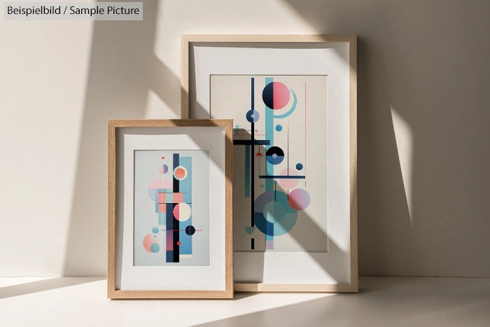 Two framed abstract prints with geometric shapes in pastel colors against a sunlit wall.