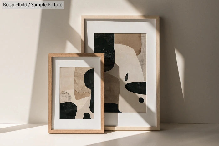 Two framed abstract paintings with black, beige, and white shapes, displayed against a sunlit beige wall.