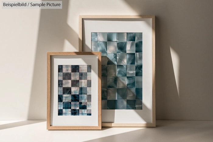 Two framed abstract artworks with checkered patterns in blue and gray tones, leaning against a sunlit wall.