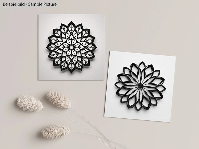 Two intricate black floral paper art designs on white backgrounds, displayed on a beige surface with decorative grasses.