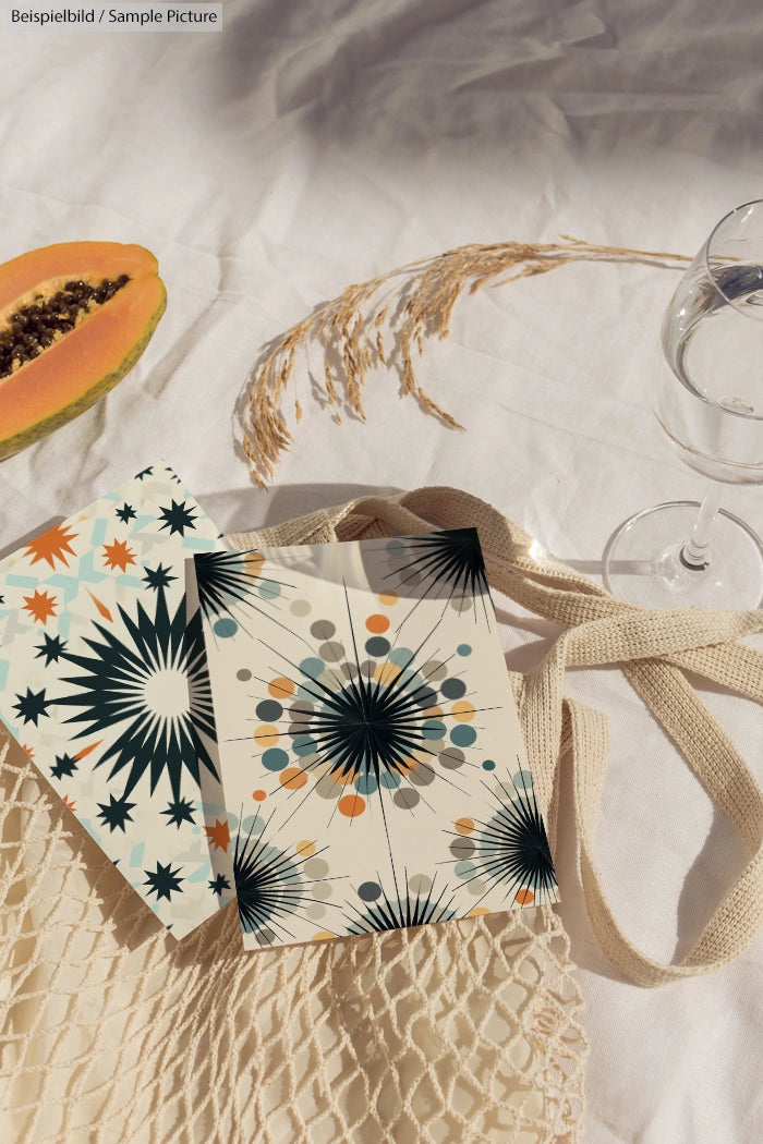 Boho-style postcards on fabric with a papaya slice, wheat sprigs, mesh bag, and a glass of water.