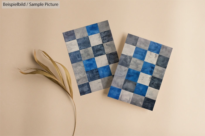 Two checkered prints with blue and gray squares on beige background, accompanied by dried plant leaves.