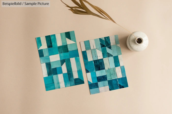 Two abstract geometric artworks with blue, teal, and gray blocks on a neutral background beside a white vase.