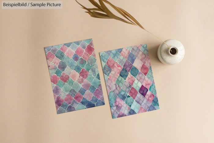 Two watercolor cards with geometric patterns next to a white vase with a dried plant on a beige surface.